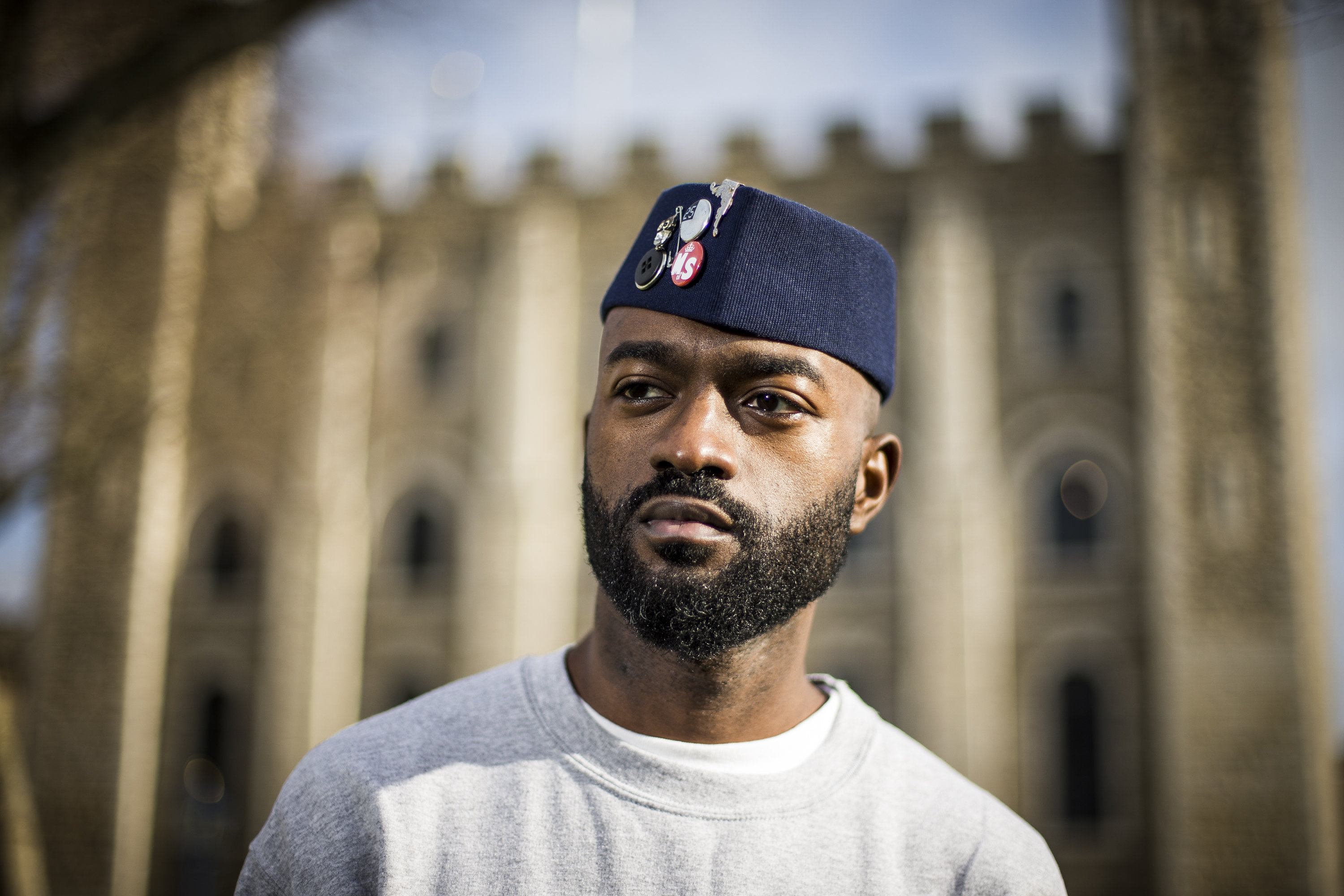 Portrait photography of Inua, wearing a grey jumper and dark blue kufi hat. Behind Inua is a faded building