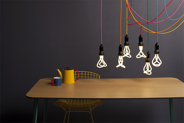 Photograph of 6 lightbulbs designed by Nicolas Roope with multi-coloured wires overhanging a wooden table with yellow kettle and blue mug.