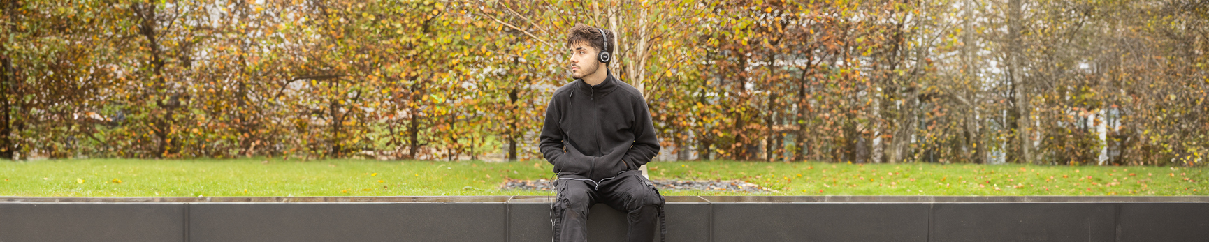 A young student wearing black clothes and headphones, sitting on a bench in front of autumn trees