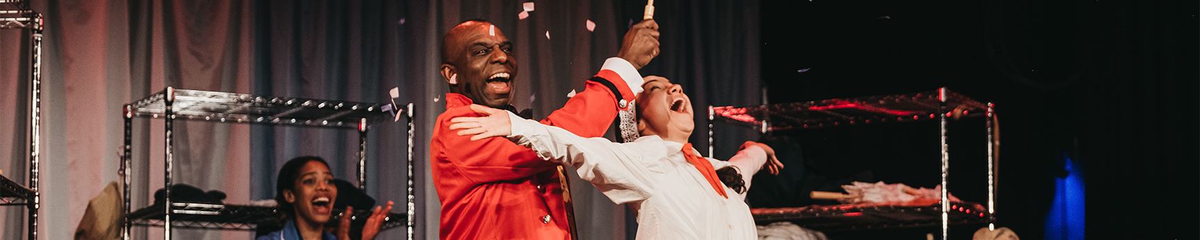 Four characters from a play, two in the foreground with an opened umbrella with confetti falling out while two in the background clap