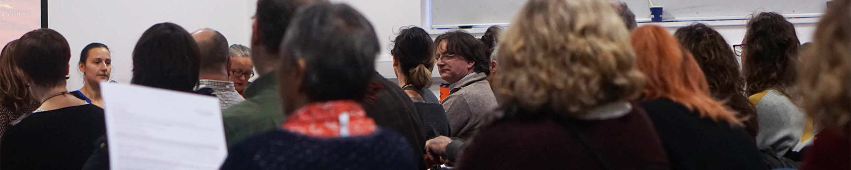 A room full of students and academics listening to a talk, some are holding paper or making notes.
