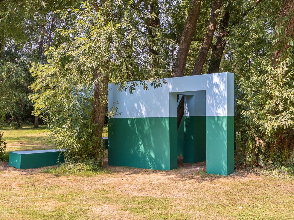 Geometric structure, half blue, half green, with doorway opening surrounded by trees