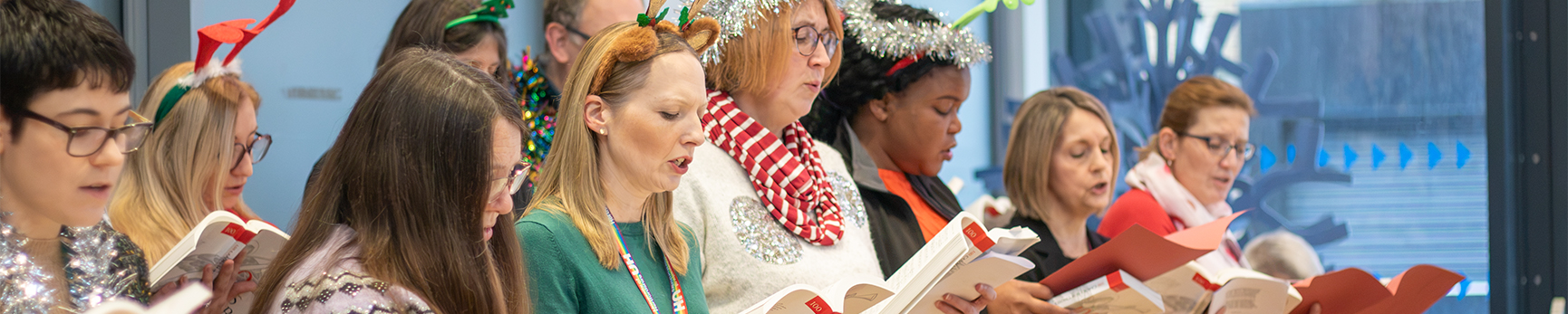 Staff and student choir singing, holding sheet music and wearing festive accessories..