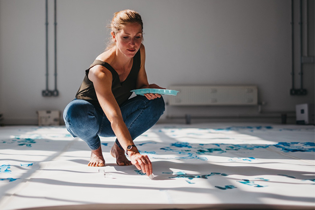 Artist creating artwork using blue inks and ice on white paper on the floor