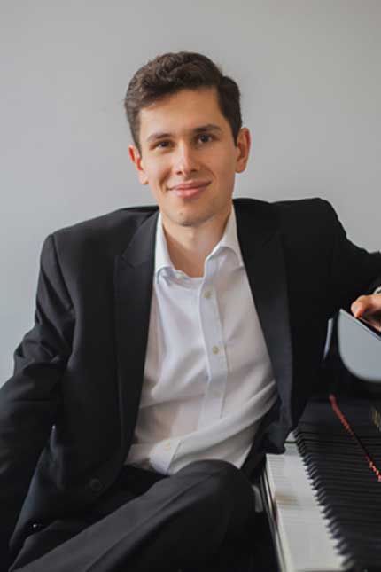 An image depicting a young, clean-shaven, white male with dark in a black suit and white shirt. He is sat down leaning on a grand piano.